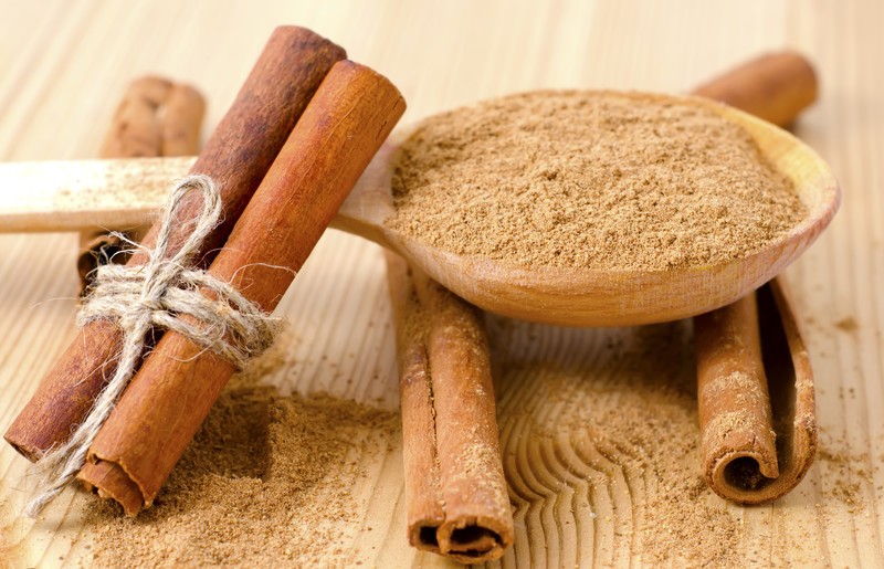 This photo shows a spoon of ground cinnamon powder with several cinnamon sticks on a wooden table.