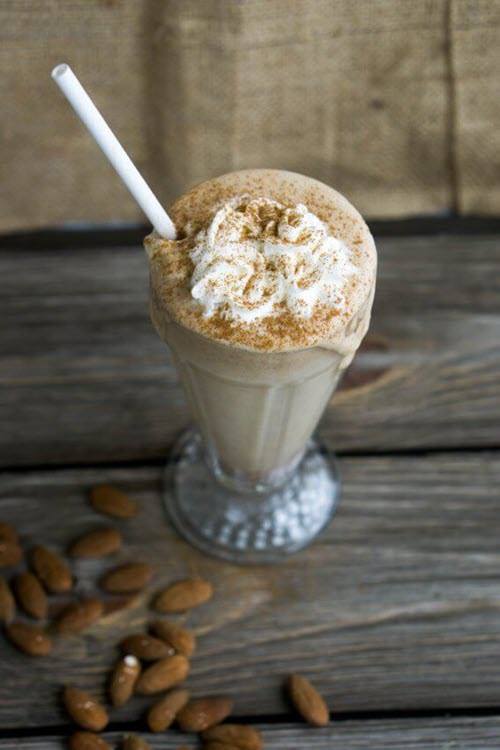 A tall glass containing a cinnamon almond butter breakfast smoothie with a straw and a pile of almonds on the table