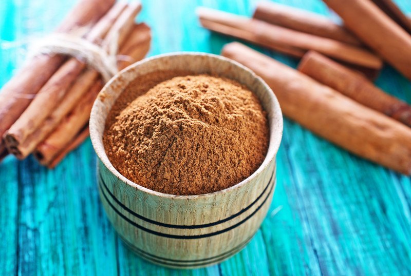 This photo shows a bowl of ground cinnamon powder and several cinnamon sticks lying on a turquoise wooden table.