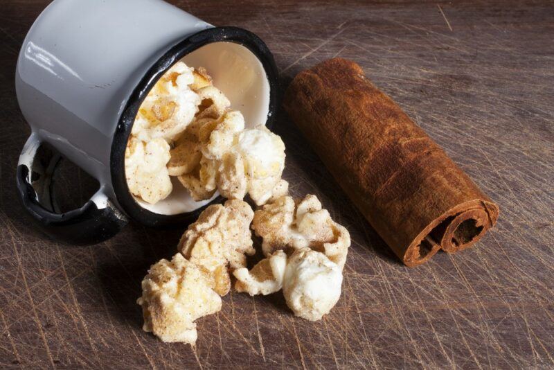 A blue mug containing cinnamon baked apple popcorn that's spilling onto the table, next to some cinnamon