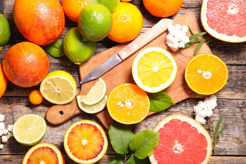 This photo shows several types of cut and uncut citrus fruits, including oranges, lemons, limes, and grapefruit on a cutting board with green leaves, all lying on a wooden table.