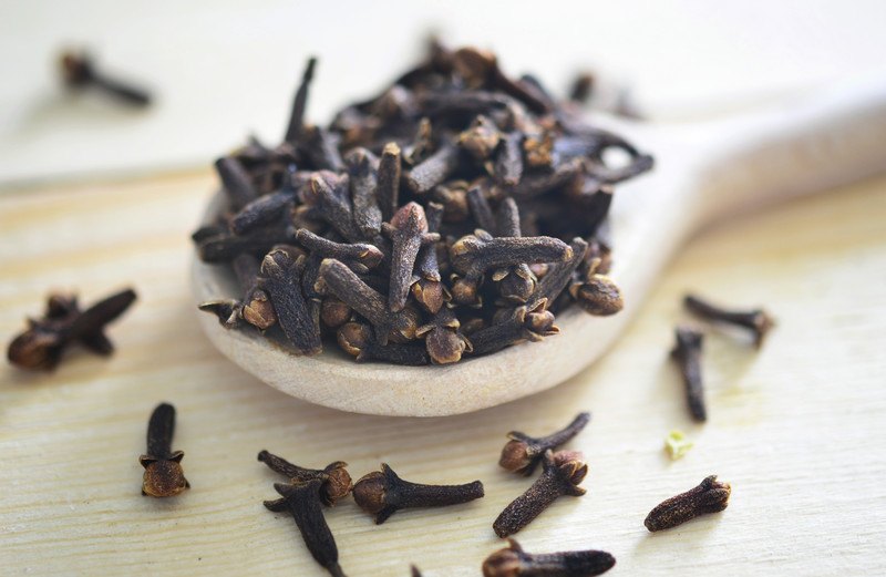 Closeup image of cloves on a wooden spoon with scattered cloves pieces around it.