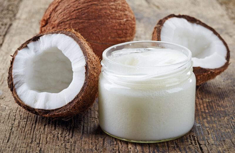 This photo shows a small open jar of coconut oil near a halved coconut and a whole coconut on a wooden surface.