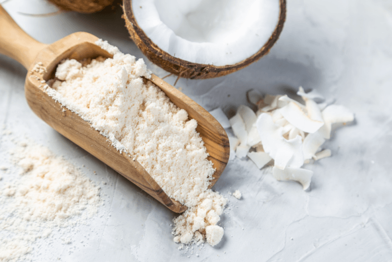 A white table with a wooden scoop of coconut flour, next to a coconut half and some shaved coconut