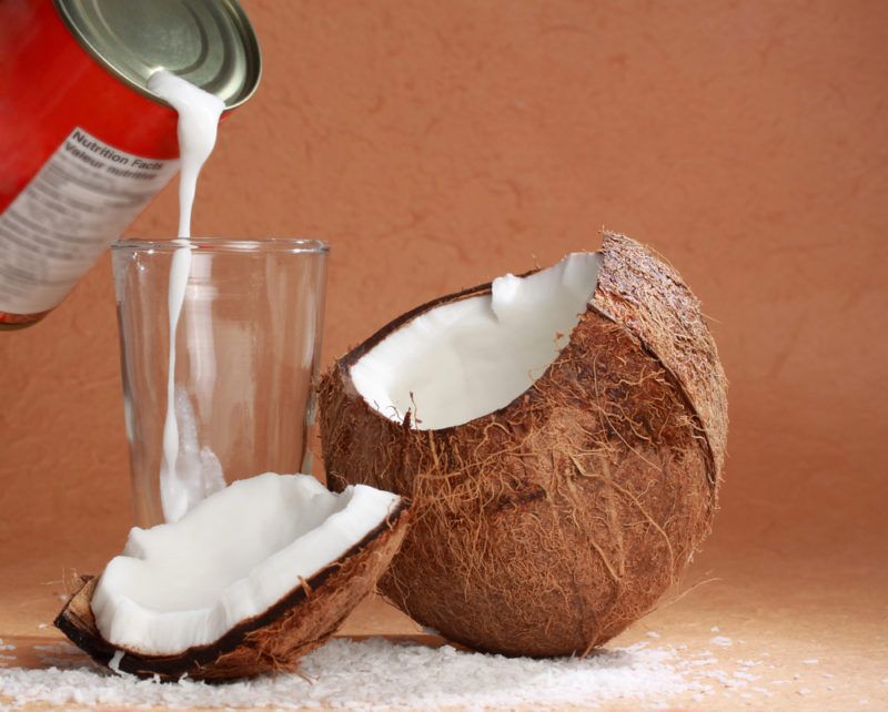 A ripe coconut, with a glass and coconut milk being poured from a can into the glass