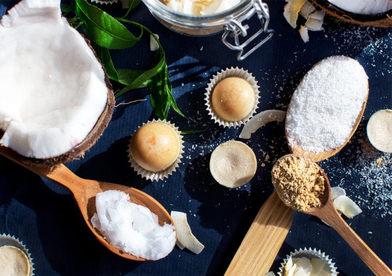 A selection of coconut oil fat bombs on a table, with all the ingredients to make them, including the coconut oil itself