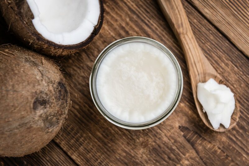 A wooden table with a whole coconut, a coconut half, a jar of coconut oil, and a spoon of coconut oil