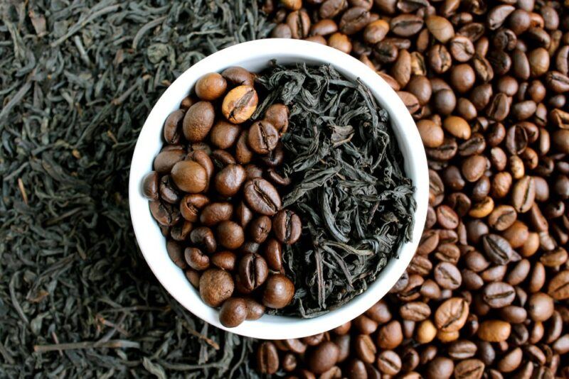 Tea leaves and coffee beans on a table with a white bowl containing the same, in contrast with each other