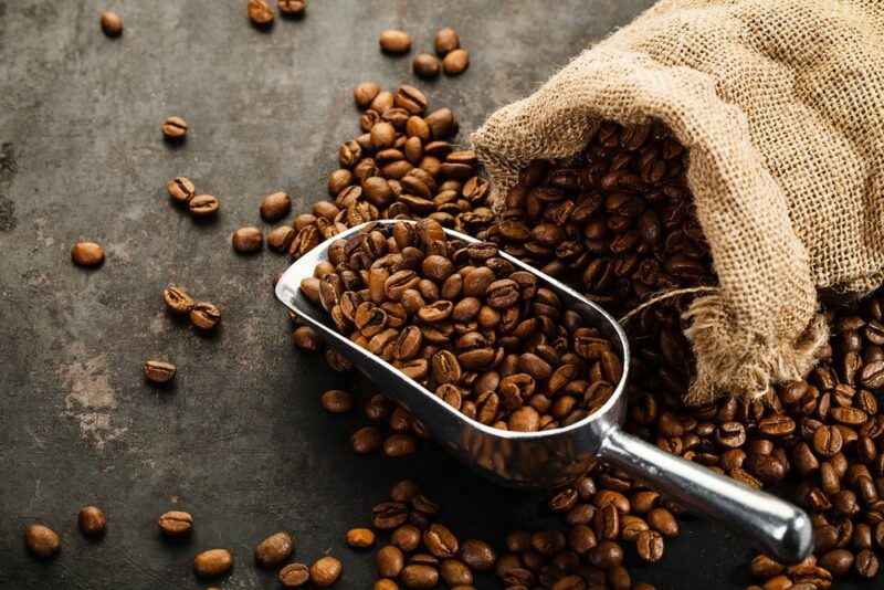 Coffee beans in a metal scoop next to a burlap sack, with beans also scattered across the table