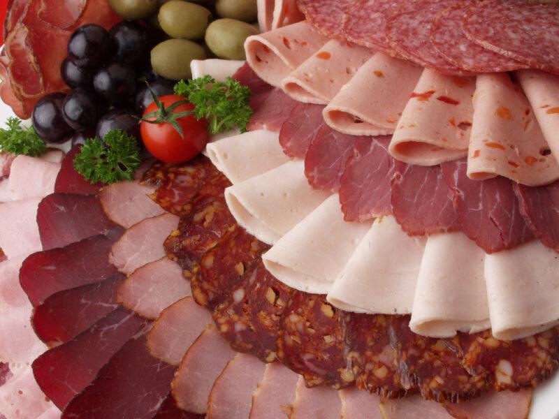 Several types of meats are layered and arranged in rows on a cold cut tray with olives, parsley, and a cherry tomato.