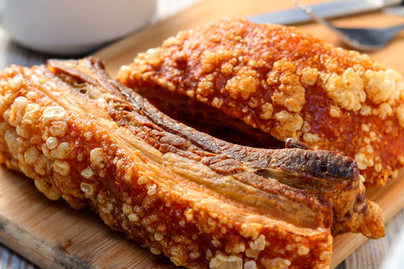 A wooden board with two cooked pieces of pork belly, where one is on its side so you can see the different layers