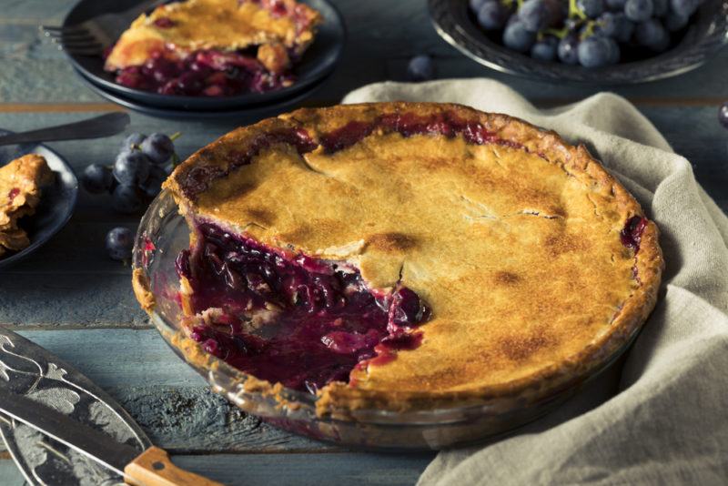 A metal dish with a concord grape pie, where one piece has been cut out and is on a black plate in the background