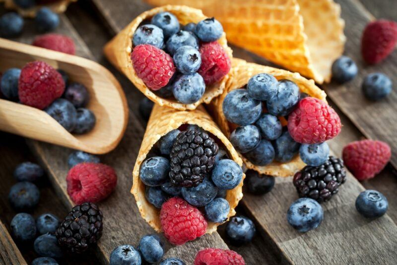 Waffle picnic cones filled with fresh berries on a wooden table, next to a wooden scoop with more berries