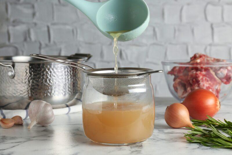 consomme a green scoop ladle pouring broth onto a metal strainer that is resting on a mason jar, at the back are some fresh vegetables and an aluminum pot