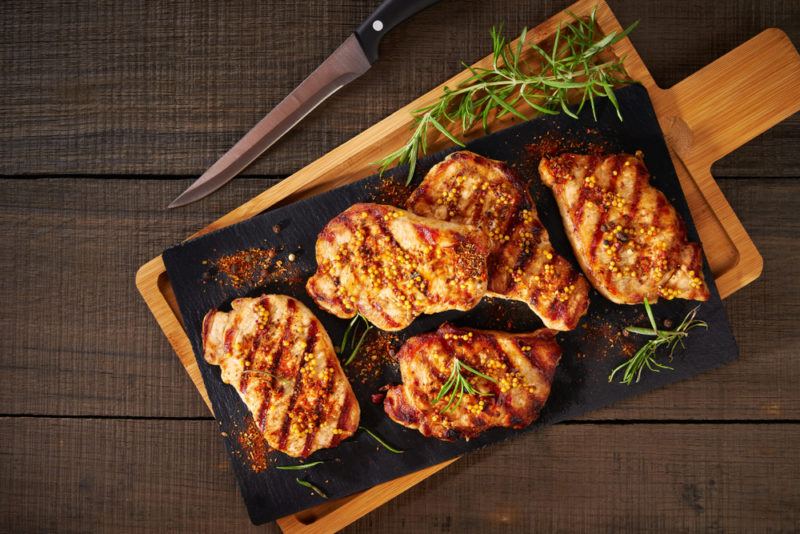 A black slate containing grilled chicken thighs and some herbs, on a wooden board that is on a wooden table