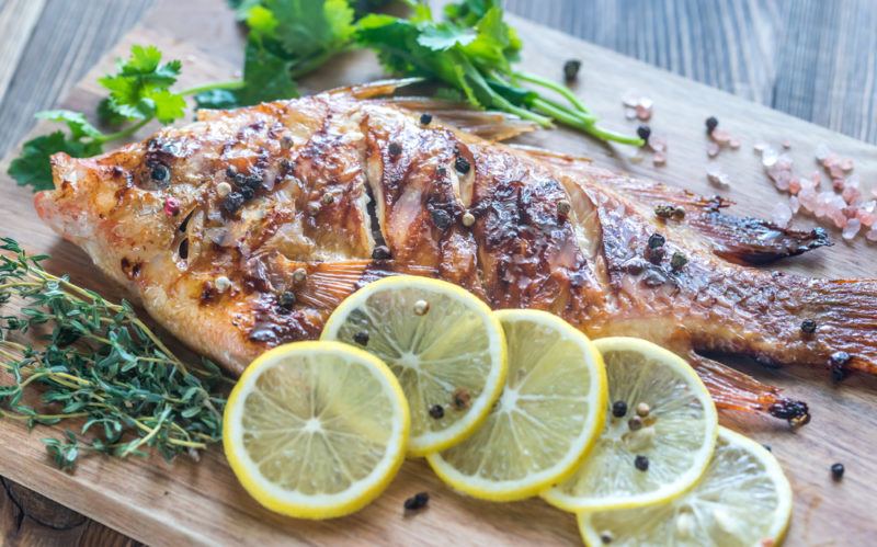 A wooden board with cooked fish, herbs, and sliced lemons