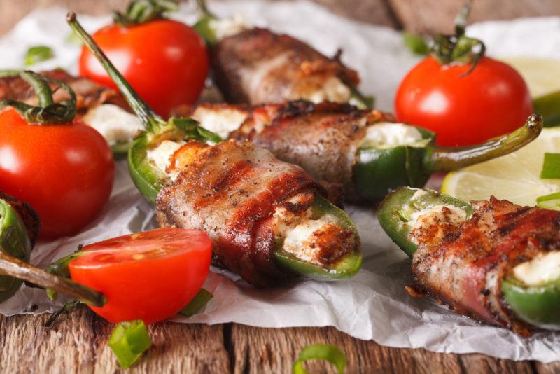 A selection of jalapeno poppers wrapped in bacon, next to fresh red tomatoes