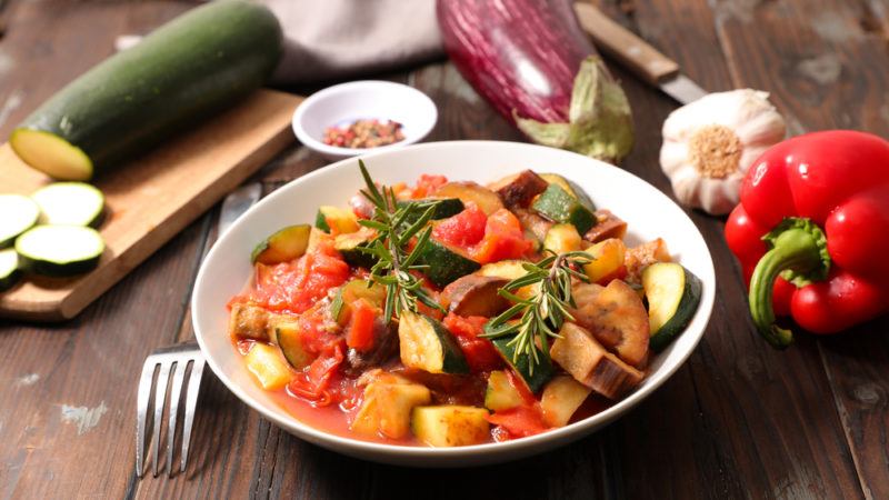 A bowl of cooked ratatouille with the various raw vegetables in the background