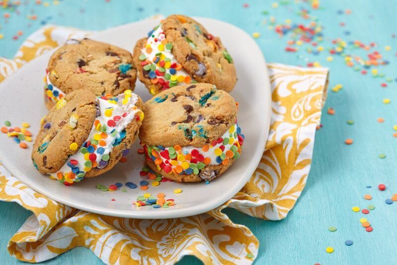 A light blue table scattered with funfetti, with a yellow piece of cloth and a plate that holds four chocolate chip cookie and funfetti sandwiches