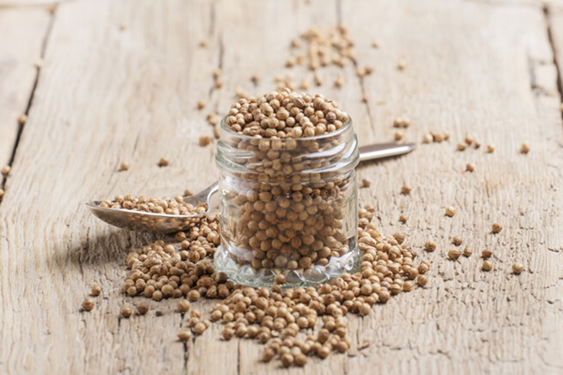 small spice jar full of coriander seeds with scattered seeds around it and a silver teaspoon, on an aged-wooden table