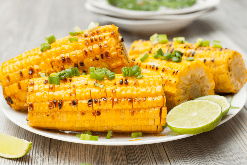 A white plate with three grilled ears of corn garnished with spring onions, plus a couple of slices of lime