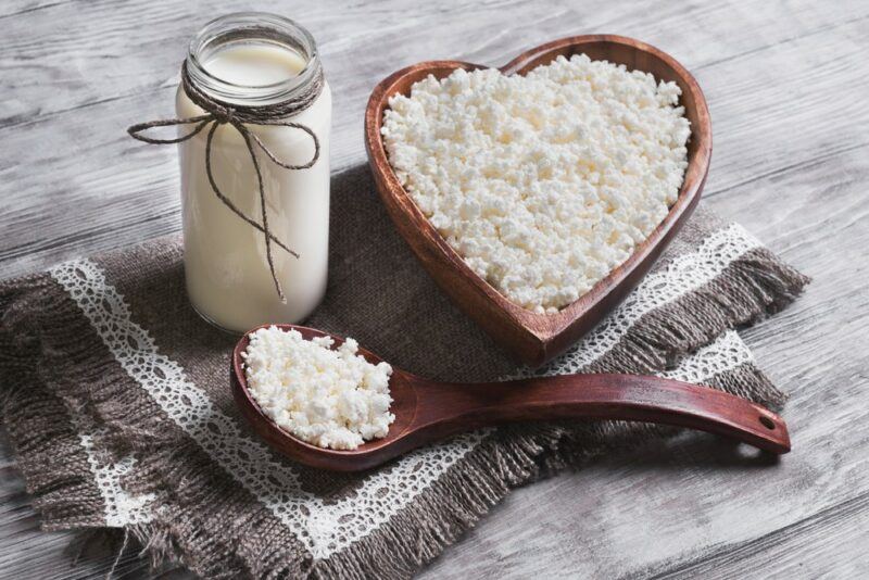 A heart shaped brown dish filled with cottage cheese, next to a spoon of the cottage cheese and a small bottle of milk