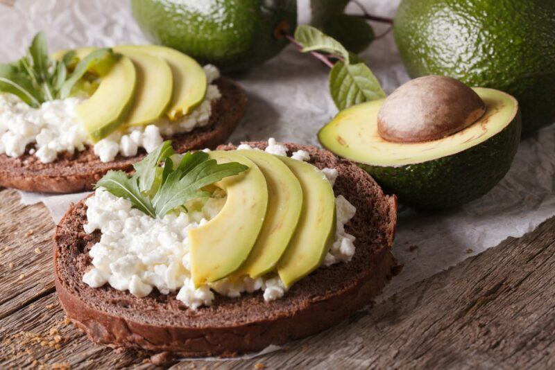 Two pieces of rye bread with cottage cheese and avocados, in front of avocados