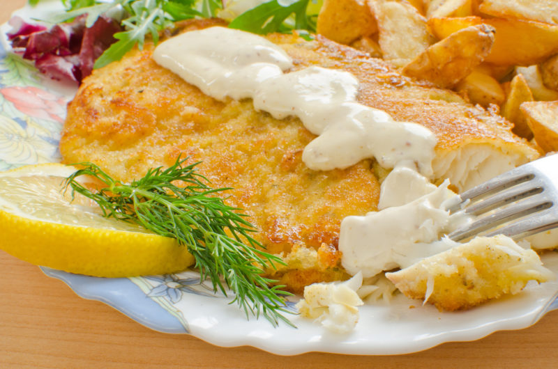 A piece of crumbed and cooked tilapia on a plate with tartare sauce, lemons, and a fork