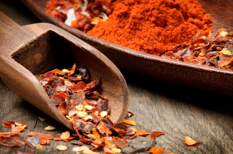 Crushed red pepper on a wooden scoop with a dish of powdered and crushed red pepper in a wooden oblong dish at the back.