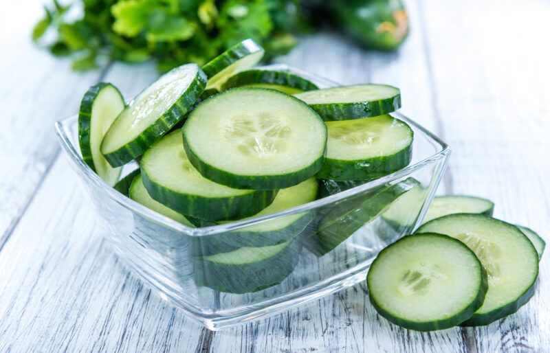 A square transparent dish holds several cucumber slices next to a few loose cucumber slices on a wooden surface.