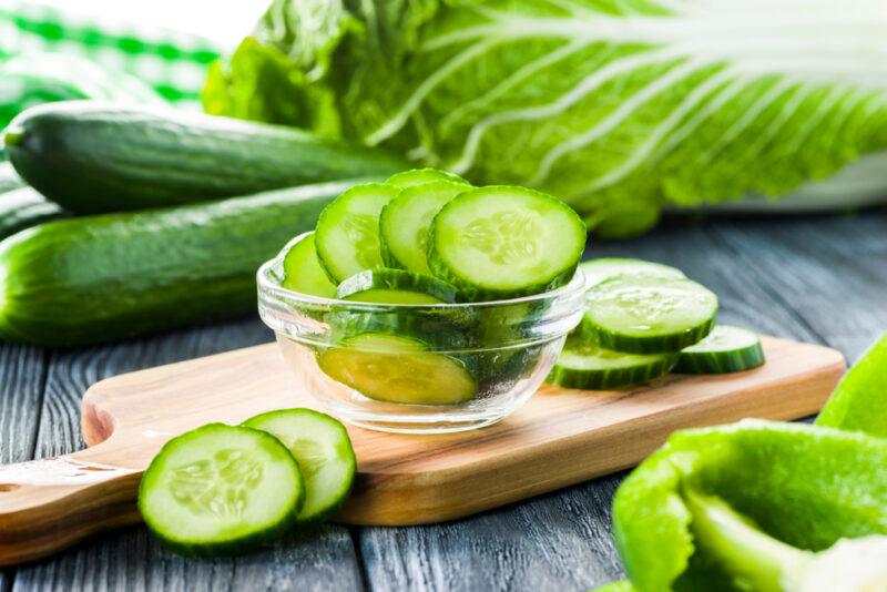 Several green cucumber slices rest in a clear bowl and on a cutting board on a dark wooden surface near full cucumbers and other green vegetables.