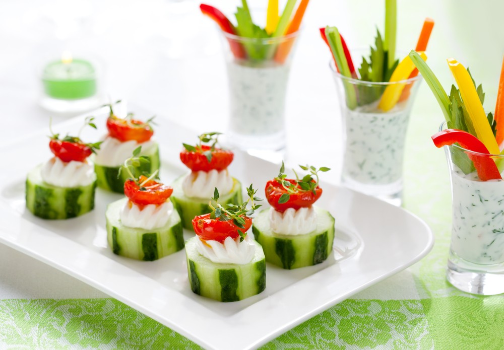 A white plate with eight pieces of sliced cucumber, with some type of cheese and cherry tomatoes, next to shot glasses filled with dip and sliced veggies