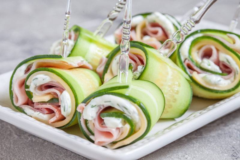 A white plate with rolls of cucumber, cream cheese and meat