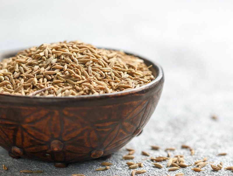 closeup image of brown ceramic bowl full of cumin seeds with a few loose cumin seeds around it