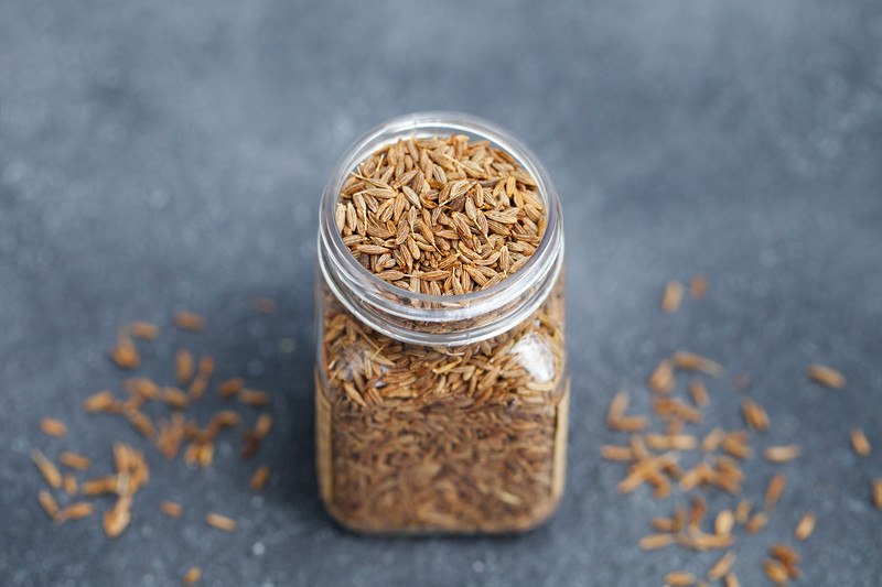 closeup image of a small jar full of cumin seeds with loose cumin seeds around it