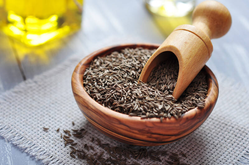 Wooden bowl full of cumin seeds with wooden scoop, resting on a cloth table mat on a wooden table. 