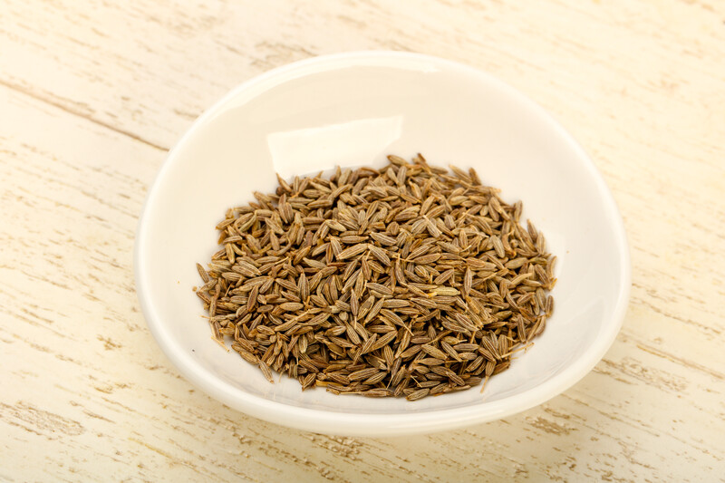 While odd-shaped ceramic bowl full of cumin seeds resting on top of a white wooden surface.