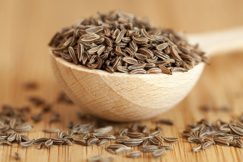 a wooden bowl with cumin seeds on a wooden table with scattered cumin seeds around it.