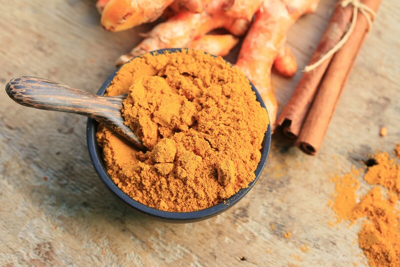 top view image of a bowl full of curry powder with a wooden spoon, resting on a wood table with fresh turmeric and cinnamon sticks