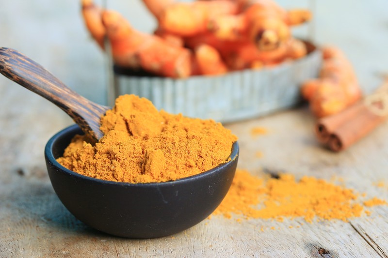 Black stone bowl full of curry powder with wooden spoon, with loose curry powder behind the bowl, and blurred image of fresh turmeric and cinnamon sticks at the back.