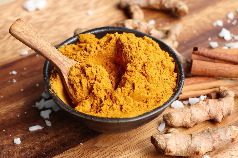 black ceramic bowl full of curry powder with a wooden spoon, resting on top of a wooden surface, with fresh turmeric and cinnamon sticks around it