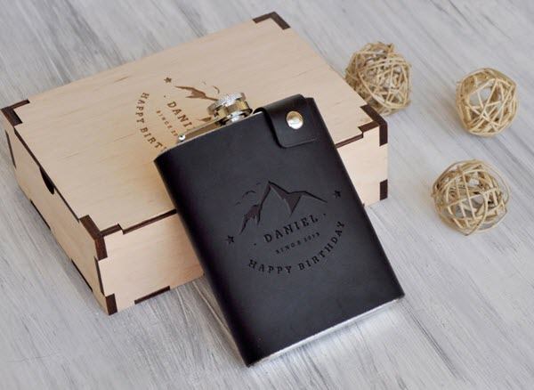Customized-Leather-Flask | Food For Net A dark leather flask with a mountain and text resting against a box with a similar engraving.