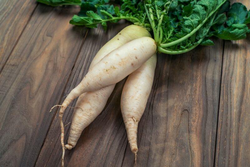 A wooden table with three white daikon radishes. Some of their greens can be seen at the top of the image too.