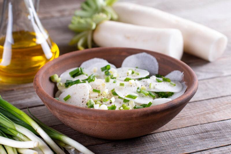 on a wooden surface is a wooden bowl full of Daikon radish salad with chopped chives on top, around it is a couple of whole radish, jar of olive oil, and bunch of chives