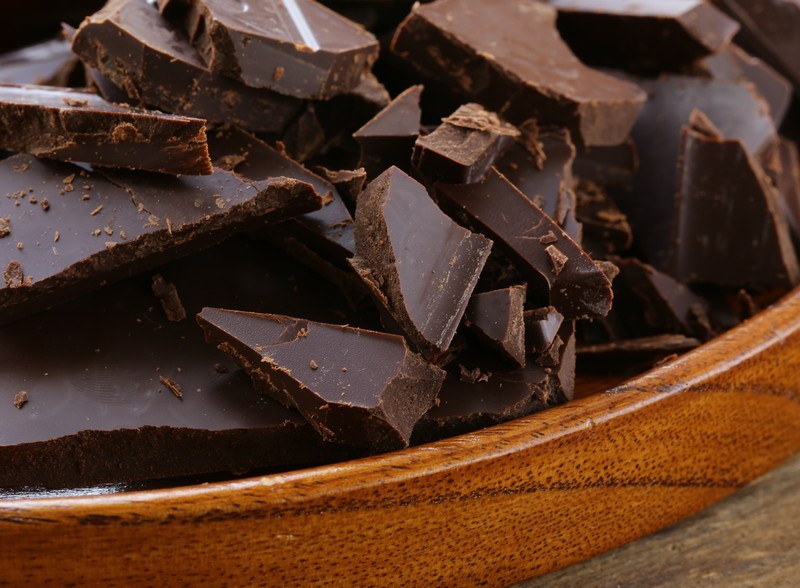 A wooden bowl filled with dark chocolate chunks rests on a wooden tabletop.