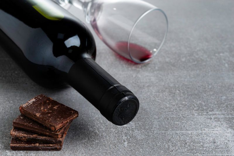 A small pile of dark chocolate, next to a bottle of red wine and a glass with a tiny bit of red wine