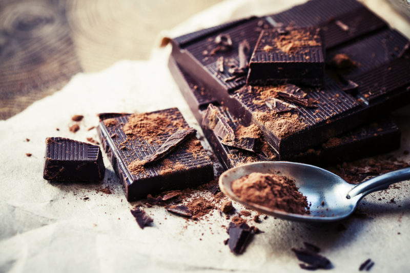 Squares of dark chocolate on paper, along with a metal spoon that has dark chocolate shavings