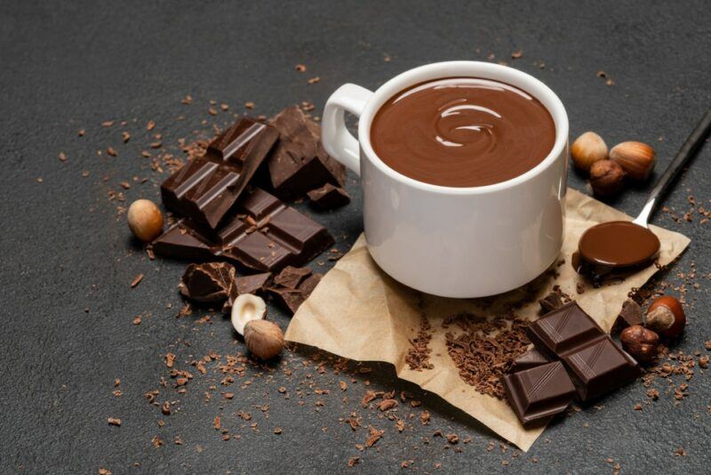 A dark table with a white mug of hot chocolate, with various pieces of dark chocolate