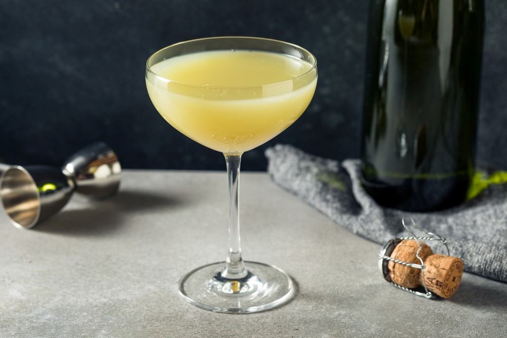 A single coupe glass on a table containing a death in the afternoon cocktail with a cocktail jigger and a bottle in the background