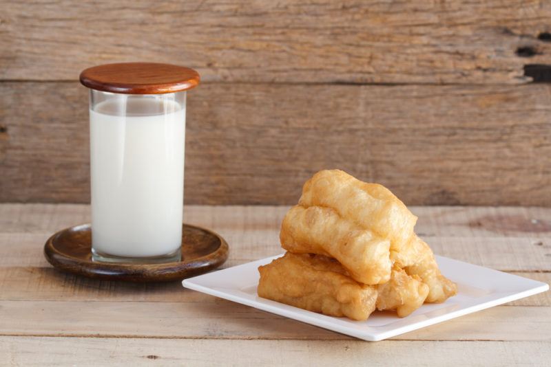 A white plate with deep fried dough sticks, next to a glass of soy milk